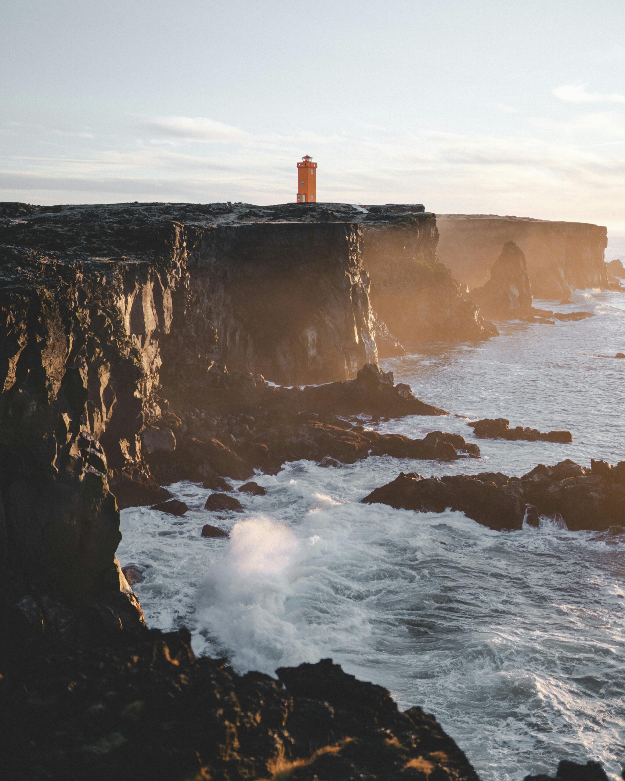 Stunning view of a lighthouse on Iceland's rugged coastline during sunset, showcasing dramatic cliffs and ocean waves.