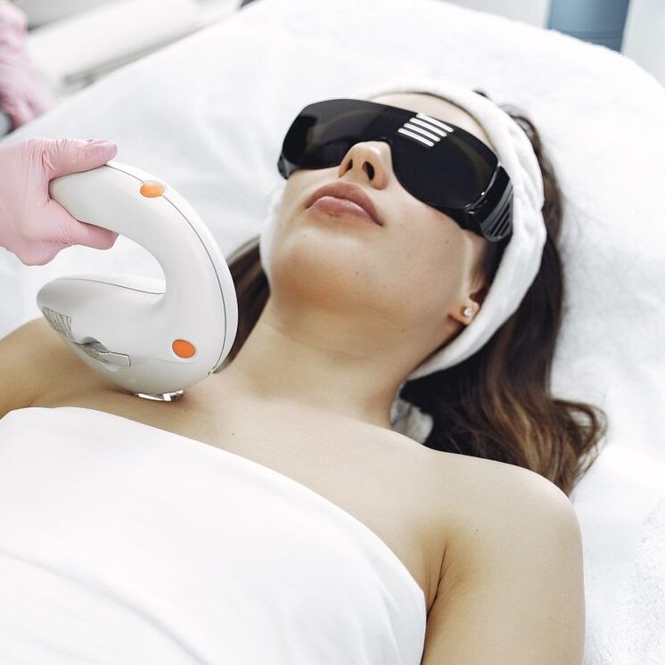 Crop faceless woman in medical uniform and gloves using laser tool on young woman wearing safety glasses and towel lying on couch in hospital