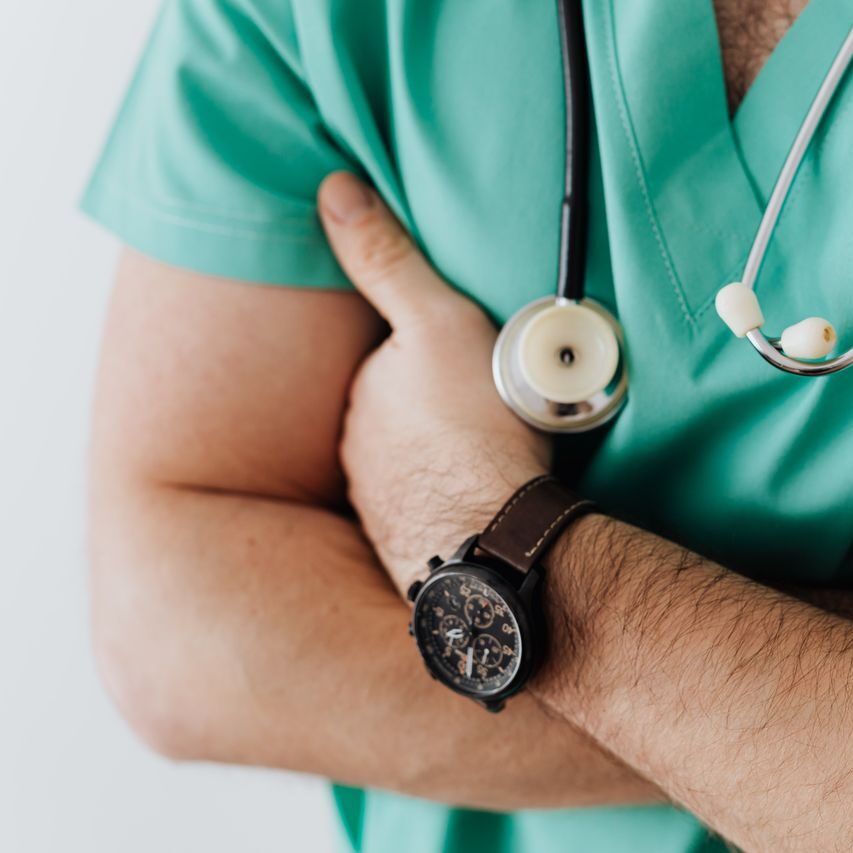 Unrecognizable man wearing medical uniform with stethoscope and pens standing in hospital with crossed arms during work in day time